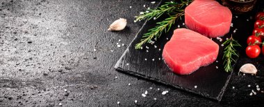 Raw tuna steak on a stone board with rosemary, spices and tomatoes. On a black background. High quality photo