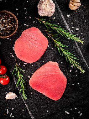 Raw tuna steak on a stone board with rosemary, spices and tomatoes. On a black background. High quality photo