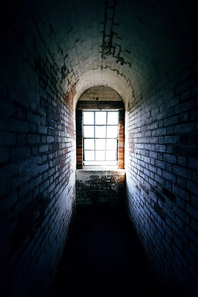 端に窓を通して光が輝いている不気味な狭い古いレンガ造りの廊下 — ストック写真
