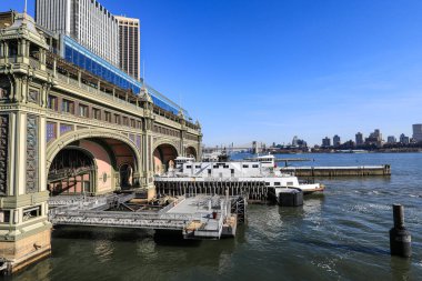 New York feribot terminalinin hareketli bir görüntüsü, feribotlar kenetlendi ve yolcuların şehir siluetinin arka planına binişi. Sahne, NYC 'nin liman taşımacılığının enerjisini ve feribotların şehir içi ulaşımdaki hayati rolünü yakalıyor.