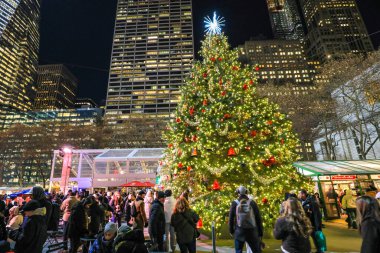 Ziyaretçiler ve turistler, New York 'taki Bryant Park Noel Pazarı' ndaki Noel ağacının etrafında toplandılar. Yanıp sönen ışıklar, tatil tezgahları ve peyzaj bahçeleriyle çevrili sahne, kış mevsiminin neşeli ruhunu yakalıyor..
