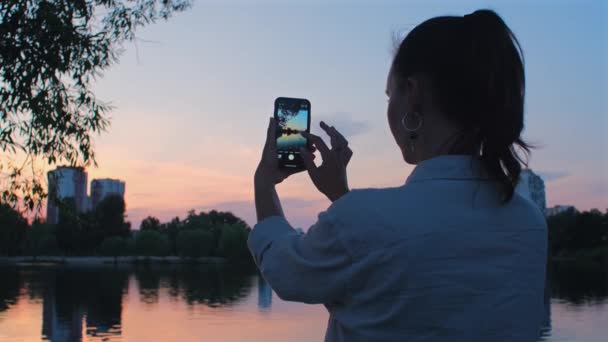 女人用手机给莱克拍照 美丽的女孩在手机上拍下了夏夜落日的照片和录像 — 图库视频影像
