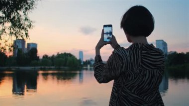 Kadın cep telefonuyla gölün fotoğrafını çekiyor. Güzel bir kız, yaz akşamında gün batımının videosunu cep telefonuyla çeker..