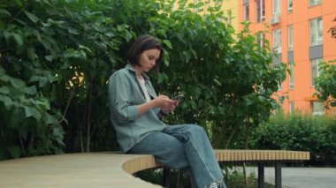 Cep telefonu kullanan genç bir kadın. İnternette gezinen akıllı telefonlu bir kız dışarıda oturuyor. Güzel esmer, sosyal medyada gezinen, mesajlaşan ya da internetten alan biri. Yüksek kalite 4k görüntü