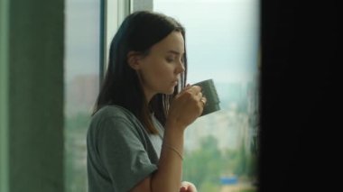 Pencerenin yanında kahve içen bir kadın. Çekici bir kadın evde dinleniyor sıcak bir içecek içiyor ve şehir manzarasına bakıyor. Evde sakin ve düşünceli bir sabah. Yüksek kalite 4k görüntü