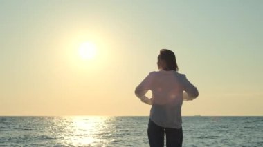 Genç kadın deniz kenarında keyifli bir sabah geçirirken ellerini kaldırıyor. Sahilde duran ve güneşin doğuşunu izleyen bir kız. Seyahat yaşam tarzı konsepti. Yüksek kalite 4k görüntü
