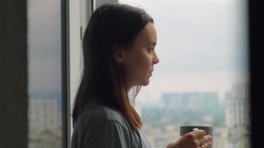 Pencerenin yanında bir fincan kahvesi olan kadın. Çekici bir kadın evde dinleniyor sıcak bir içecek içiyor ve şehir manzarasına bakıyor. Sıcak ve sakin bir sabah. Yüksek kalite 4k görüntü