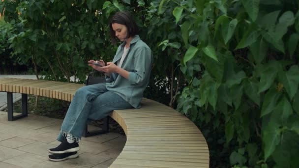 用手机的年轻女人女孩手持智能手机 坐在室外浏览互联网 可爱的黑发卷轴社交媒体饲料 发短信或网上购买 高质量的4K镜头 — 图库视频影像