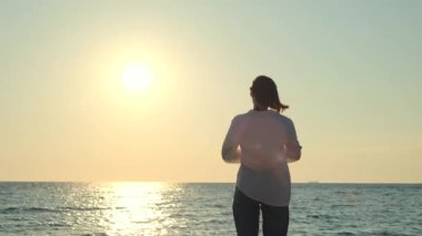 Genç bayan deniz kenarında sabahın tadını çıkarıyor. Sahilde duran ve güneşin doğuşunu izleyen bir kız. Seyahat yaşam tarzı konsepti. Yüksek kalite 4k görüntü