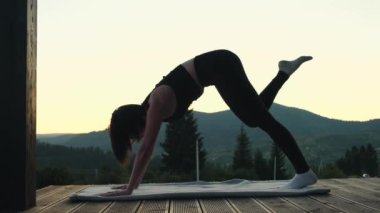 Ahşap terasta dağ manzaralı yoga yapan genç bir kadın. Kadın silueti meditasyon yapıyor ve alacakaranlıkta yoga pozları veriyor. Doğa manzarası. Yüksek kalite 4k görüntü