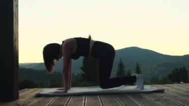 Genç atletik kadın dağ manzaralı ahşap terasta yoga yapıyor. Kadın silueti meditasyon yapıyor ve alacakaranlıkta yoga pozları veriyor. Doğa manzarası. Yüksek kalite 4k görüntü
