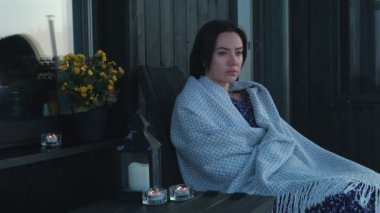 Kır evinin terasında oturan üzgün genç kadın. Sıcak battaniyeye sarılı depresif mutsuz kız doğa manzarasına bakıyor. Yüksek kalite 4k görüntü
