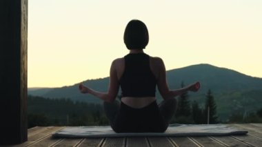Genç bir kadın dağ manzaralı ahşap terasta meditasyon yapıyor. Kadın silueti alacakaranlıkta yoga pozu veriyor. Doğa manzarası işte. Yüksek kalite 4k görüntü