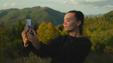 Dağ zirvesinde cep telefonuyla selfie çeken genç bir kadın. Mutlu kız cep telefonu kamerasıyla fotoğraf çekiyor, manzara manzarası arka planda. Yüksek kalite 4k görüntü