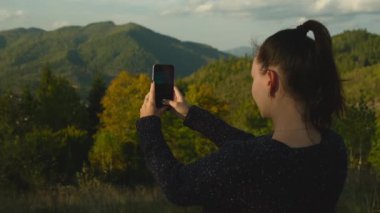 Mutlu kız cep telefonu kamerasıyla dağların fotoğraflarını çekiyor. Genç bir kadın dağın zirvesinde duruyor ve cep telefonu cihazıyla manzaranın fotoğrafını çekiyor. Yüksek kalite 4k görüntü