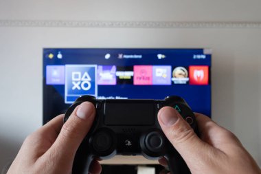 Bir erkek el, arka plandaki akıllı bir TV 'de arayüz içinde bir video oyunu arayan bir Playstation 4 denetleyicisini tutuyor.