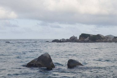 Güzel Karayipler deniz mavisi manzarası, büyük kayalar ve arka planda bir kayanın tepesinde oturan bir adam.