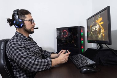 Young latin man gamer with long hair glasses and headsets holding a controller on his hands playing call of duty black ops 6 on a gamer pc clipart