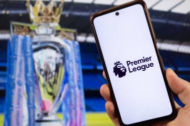 BOGOTA, COLOMBIA - 05 Mart 2025 Premier Lig futbol logosuna sahip akıllı telefonu beyaz bir arkaplanda tutan ve arka planda bulanık bir premier lig kupası