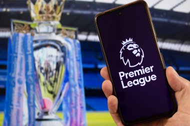 BOGOTA, COLOMBIA - Premier Lig futbol aslanı logosuna sahip bir akıllı telefon ve arka planda bulanık Premier Lig kupası