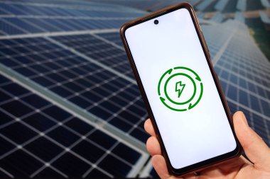 Arka planda bulanık güneş paneli olan temiz enerji logosuna sahip akıllı telefonu tutan bir erkek el.