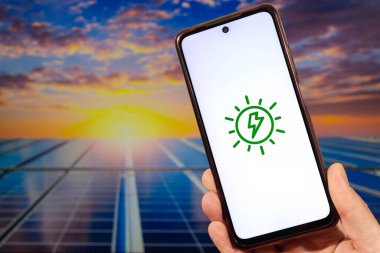 Sağ eliyle akıllı telefonu tutan bir erkek el ve arka planda bulanık güneş paneli olan bir eolik enerji logosu.