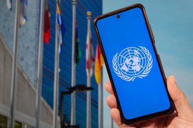 Birleşmiş Milletler ya da BM logosu olan akıllı telefonu tutan bir erkek el arka planda BM karargahını bulanıklaştırdı