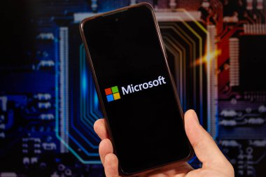 Sağ eliyle akıllı telefon tutan bir erkek ve arka planında bulanık bir anakart duvar kağıdı olan Microsoft logosu.