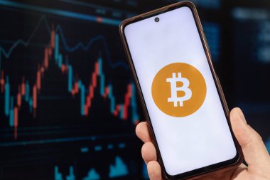 Elinde Bitcoin logosu ve arka planda bulanık bir borsa duvar kağıdı olan akıllı bir telefon tutan bir erkek.