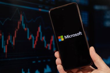 Sağ eliyle akıllı telefon tutan bir erkek ve arka planda bulanık borsa duvar kağıdı olan Microsoft logosu.