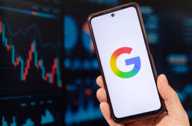 Sağ eliyle akıllı telefon tutan bir erkek ve arka planında bulanık borsa duvar kağıdı olan Google şirket logosu.