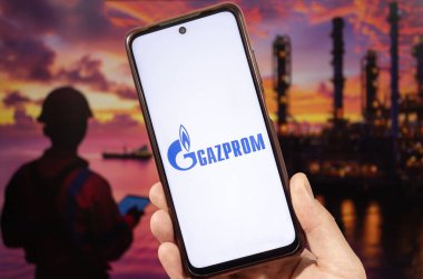 Arka planda bulanık petrol endüstrisi olan Gazprom logosu olan sağ eliyle akıllı telefon tutan bir erkek.