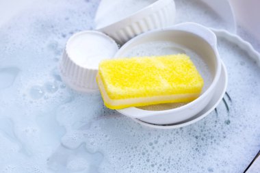Dishes and bowls with yellow sponge in water and bubbles of dishwashing liquid