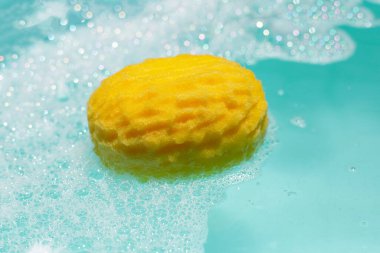 Sarı banyo süngeri sabun köpüğüyle mavi suda yüzüyor.