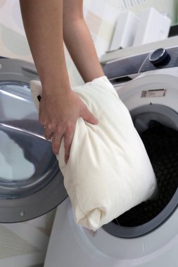 Adamın eli yastığı tutuyor ve çamaşır makinesine koyuyor..