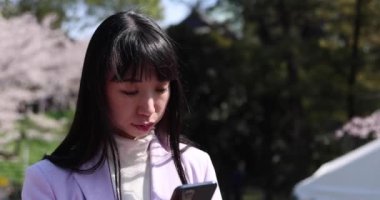 Kiraz çiçeğinin arkasında akıllı telefon kullanan Japon bir kadının portresi. Yüksek kalite 4K görüntü. Chiyoda bölgesi Tokyo 03.30.2023