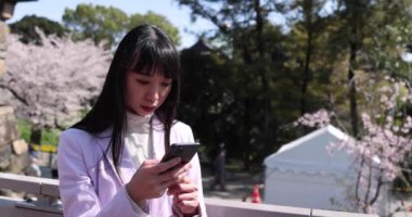 Japon bir kadının portresi kiraz çiçeğinin arkasındaki akıllı telefonu arıyor. Yüksek kalite 4K görüntü. Chiyoda bölgesi Tokyo 03.30.2023