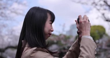 Kiraz çiçeğinin arkasında aynasız kamerayla çekim yapan Japon kadının portresi. Yüksek kalite 4K görüntü. Chiyoda bölgesi Tokyo 03.30.2023