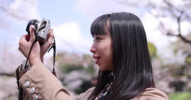 Kiraz çiçeğinin arkasında aynasız kamerayla çekim yapan Japon kadının portresi. Yüksek kalite 4K görüntü. Chiyoda bölgesi Tokyo 03.30.2023