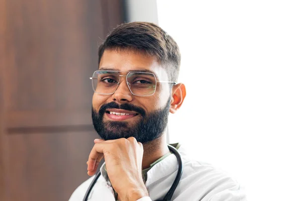Portrait of male indian doctor wearing white coat in clinic office.