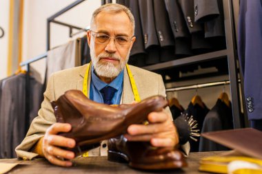 Gri sakallı yaşlı yakışıklı adam atölyede güzel bir takım elbise giyer ve el yapımı itatik ayakkabı kalitesini ölçer..