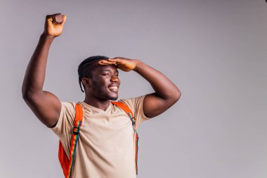 Gri arka plan stüdyosunda turuncu bir sırt çantası ve defter tutan bej tişörtlü polyetik bir adam..