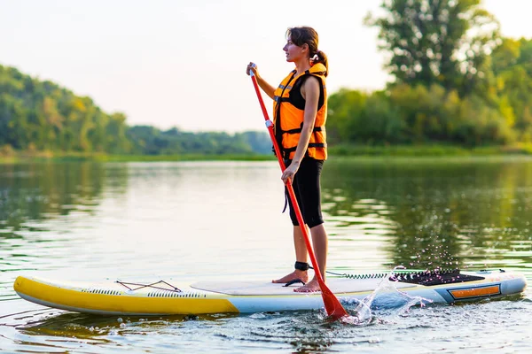 woman in life jacket at sub board at river ar evening , forest trees background .