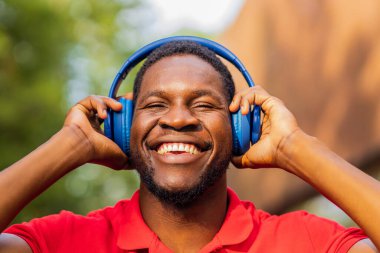 african american man in red t-shirt listening music in blue headphones and being in high spirit outdoors .