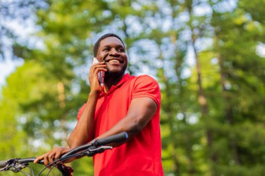 afro american man in red t-shirt walking in park with a bicycle.