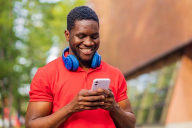 young afro american man with headphones on neck using smartphone in summer park .