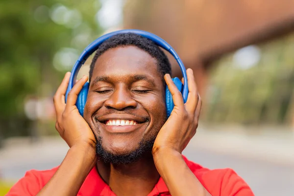 stock image african american man in red t-shirt listening music in blue headphones and being in high spirit outdoors .