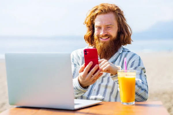 young redhaired ginger bearded man working outdoors in sea cafe with laptop an summer day.
