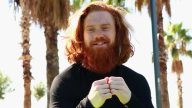 Uzun sakallı yakışıklı kızıl saçlı adam güneşli sahilde biriyle konuşurken siyah spor takım elbise giyiyor..