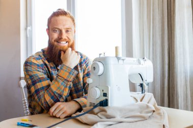 Uzun sakallı yakışıklı kızıl saçlı adam dikiş makinesinde dikiş dikiyor..